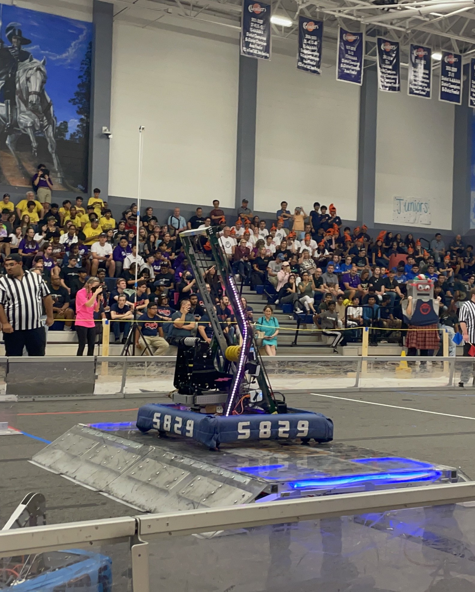 A photo of a 2023 FRC game. The Awtybots robot is on the charge station, balancing alone, while there is a crowd on the stands in the background. A large poster of a horseman and a horse is visible in the top left of the image. The photo is taken from one side of the field, looking up across the field.